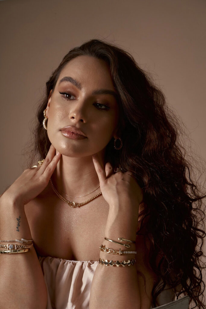 Abril Escalante a model with wavy brown hair is captured in a close-up shot. She wears gold jewelry including a necklace bracelets and earrings. Her makeup is subtle with a focus on her eyes and she has a tattoo on her left wrist.